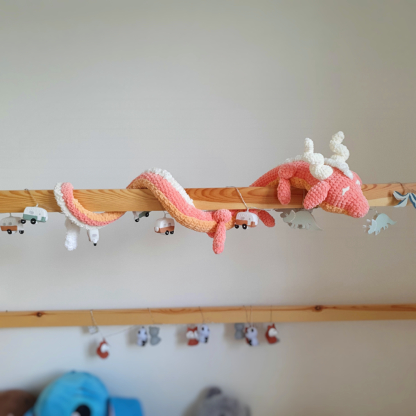 Dragon orphelin corail en laine crochetée à la main, enroulé autour d’une barre de lit – Les Doudous de Woolfy